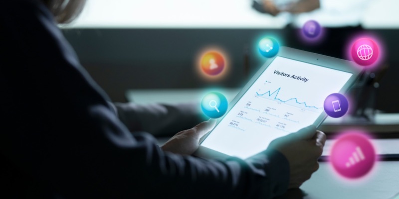 Pessoa a utilizar um tablet com gráficos de análise de dados, rodeado por ícones digitais que representam ferramentas e canais do marketing digital.