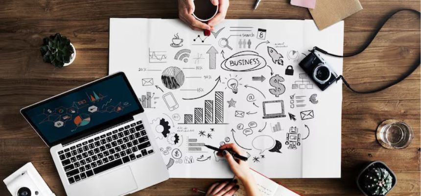 Esta fotografia em ângulo superior captura a essência  planeamento criativo, unindo o analógico ao digital através de uma mesa de madeira onde mãos humanas interagem com um laptop, câmeras e um grande mapa mental repleto de desenhos, gráficos e ideias de negócios, transmitindo uma atmosfera de colaboração, café e execução estratégica visual.