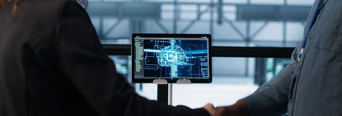 Duas pessoas vistas de perfil observam um monitor com uma imagem de cérebro digital e interface tecnológica, em ambiente de reunião, sugerindo inteligência artificial e transformação digital