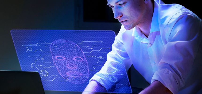 Pessoa sentada em frente a um portátil observa um ecrã holográfico azul com o desenho de um rosto digital em malha 3D e circuitos, sugerindo inteligência artificial e tecnologia de reconhecimento facial.